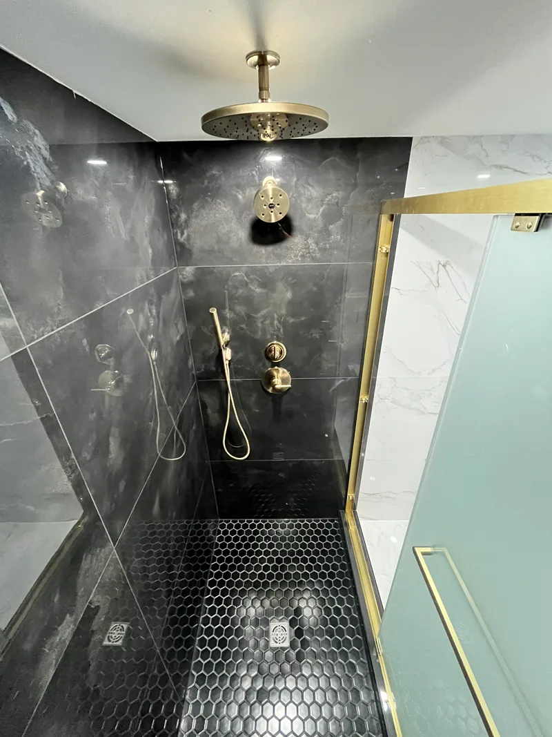Inside the finished Natick shower &mdash; black large-format porcelain walls, brass rain head and valve trim, black hex mosaic floor sloped to the drain