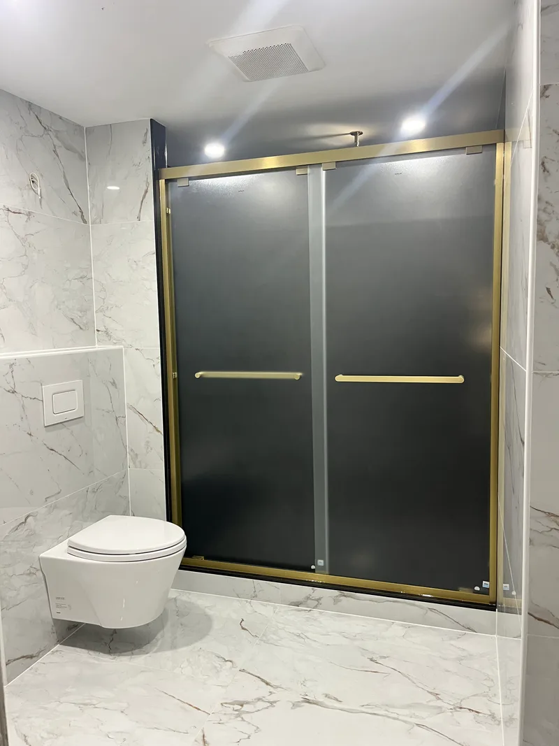 Walk-in shower entry in Natick — sliding glass in a brushed brass frame, floor tile running unbroken to the threshold
